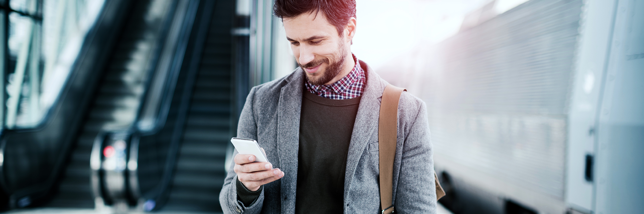 Person steht an einem Bahnhof und schaut lächelnd auf ein Smartphone.