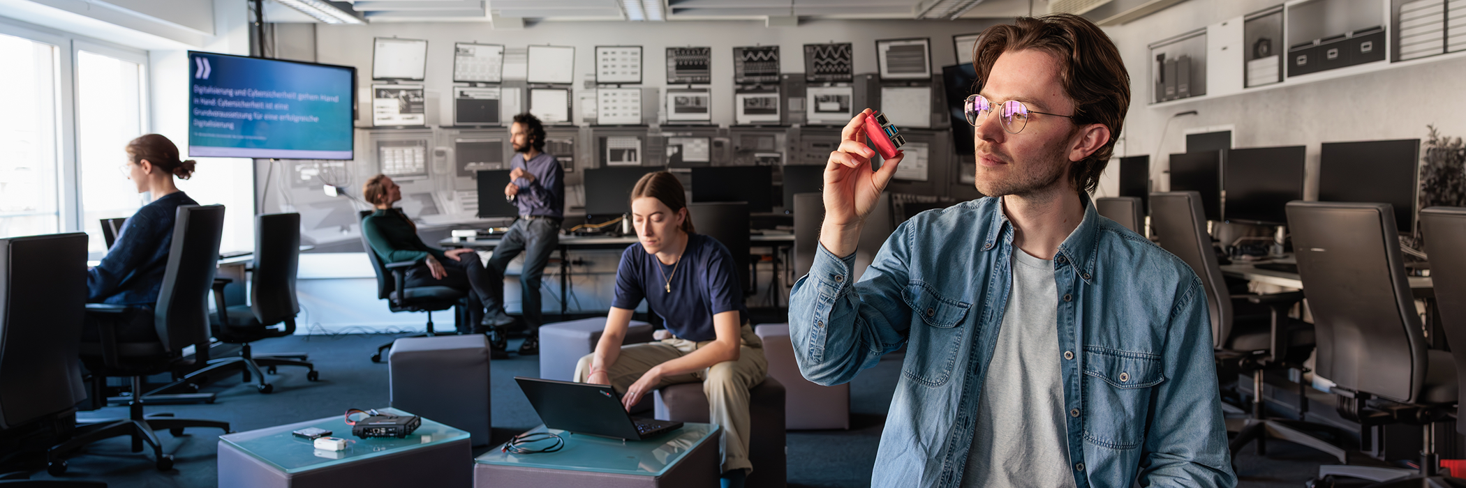 Mehrere Personen arbeiten in einem modernen IT-Schulungsraum; im Vordergrund hält jemand ein elektronisches Gerät hoch.