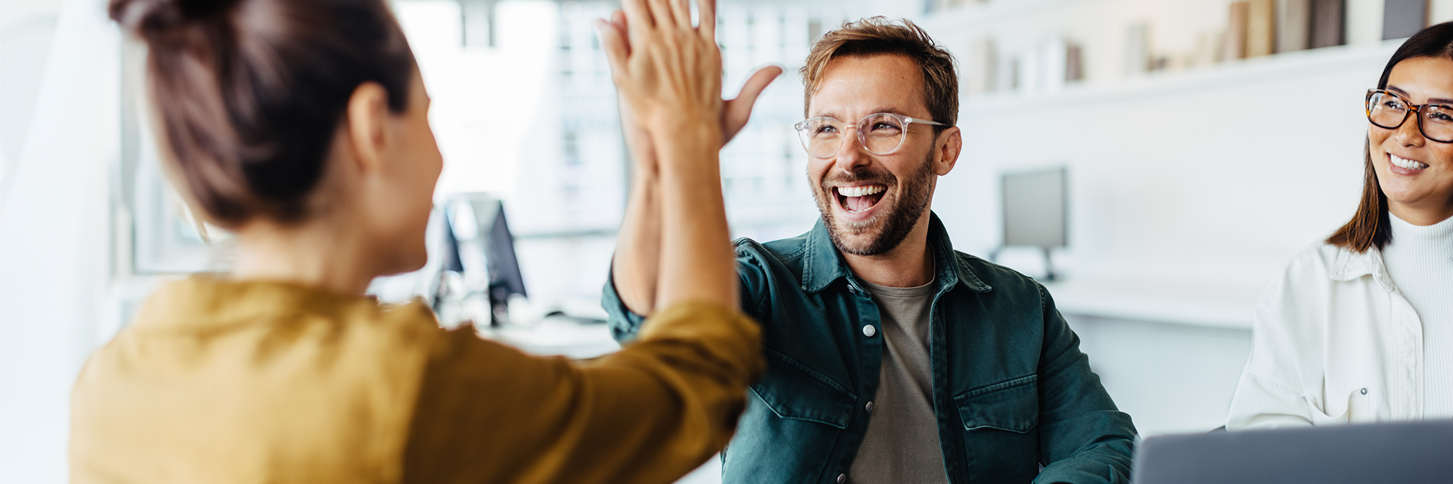 Zwei Personen geben sich ein High-Five in einem modernen Büro, dritte Person schaut lächelnd zu.