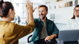 Zwei Personen geben sich ein High-Five in einem modernen Büro, dritte Person schaut lächelnd zu.