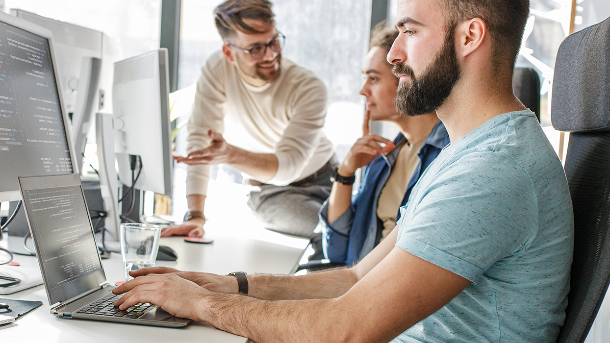Drei Personen coden und besprechen sich in einem Büro-Setting.