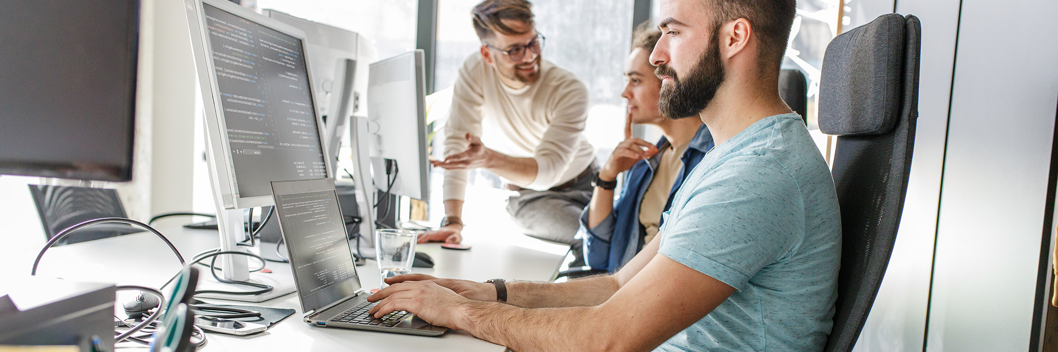 Drei Personen coden und besprechen sich in einem Büro-Setting.