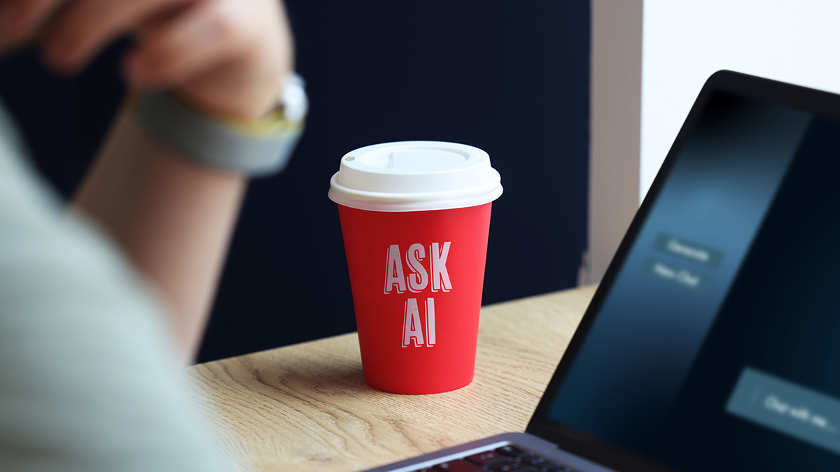 Roter Kaffeebecher mit der Aufschrift "ASK AI" neben einem Laptop auf einem Schreibtisch