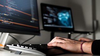 Hände tippen auf Tastatur vor zwei Monitoren mit Code-Anzeige