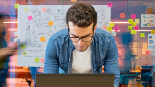 Person arbeitet konzentriert am Laptop in kreativem, farbig gestaltetem Büro mit Notizzetteln im Hintergrund.